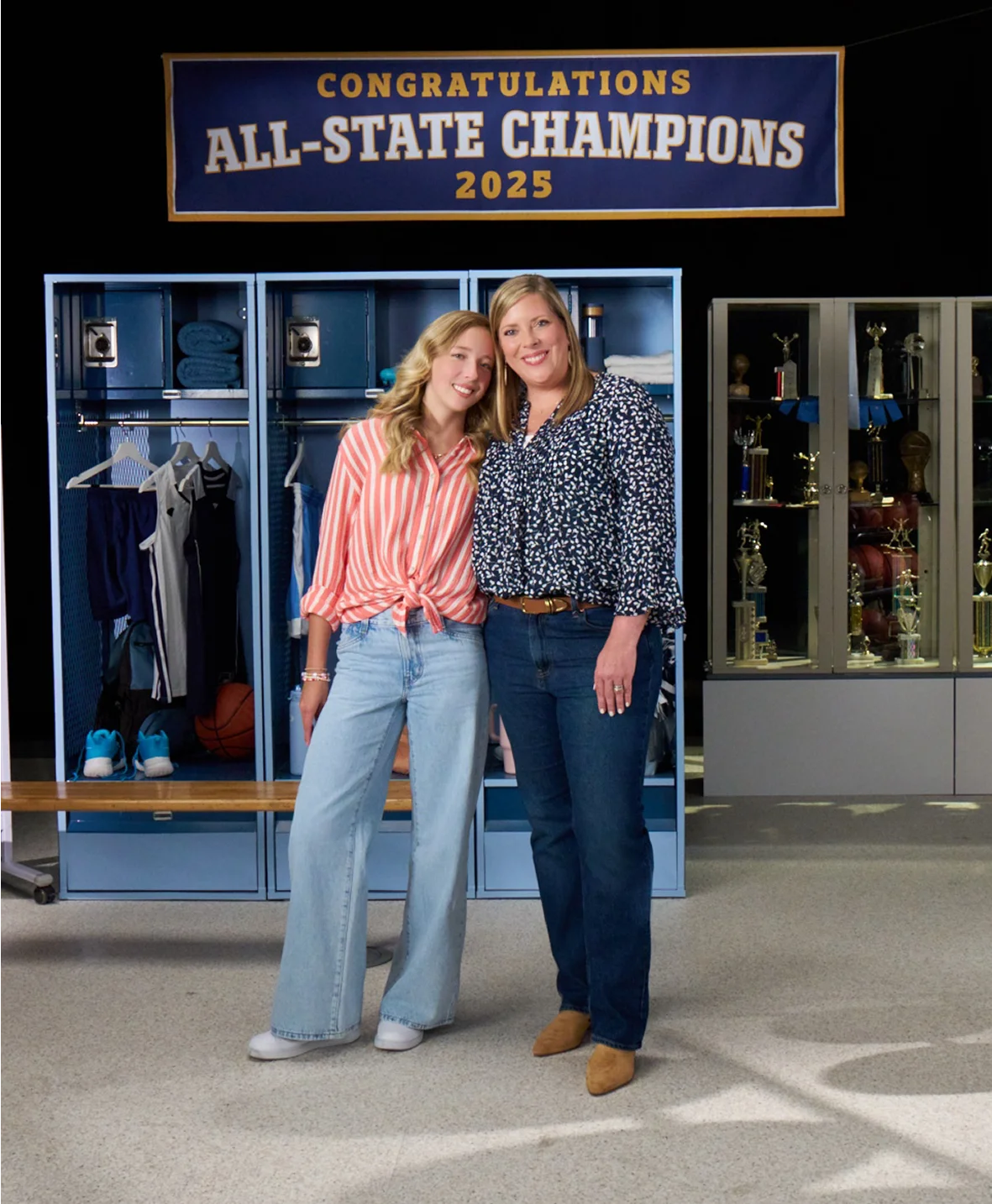 Anna and Rachael standing in front of an All-State Champions sign, lockers, a trophy case, and sports equipment, as they discuss managing eczema with Opzelura.