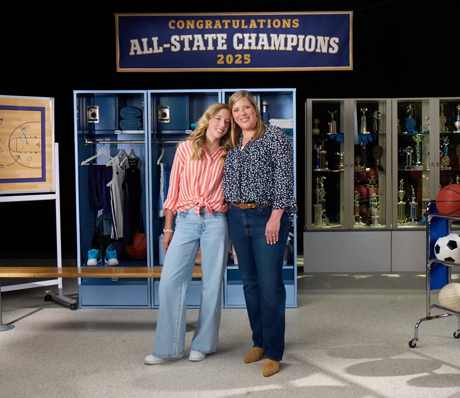 Anna and Rachael standing in front of an All-State Champions sign, lockers, a trophy case, and sports equipment, as they discuss managing eczema with Opzelura.
