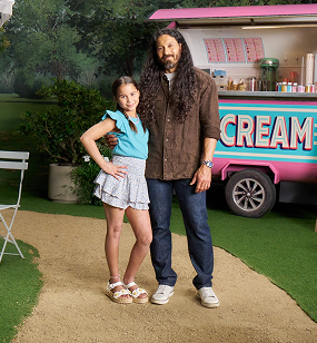 Piper and Adam standing in a park setting in front of an ice cream cart, discussing managing eczema at a young age with Opzelura.